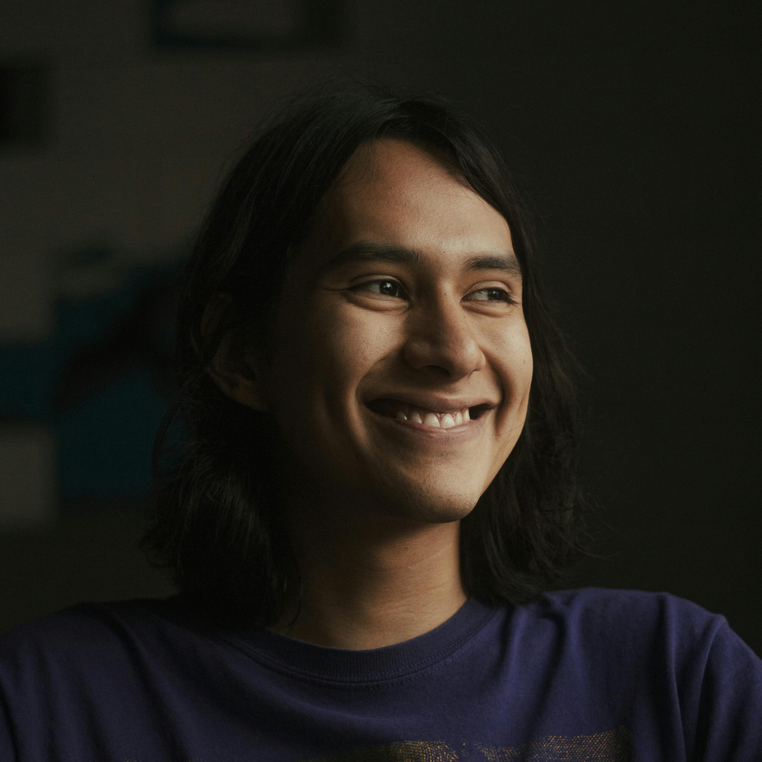 Young man with long hair smiling warmly in a dimly lit indoor setting.